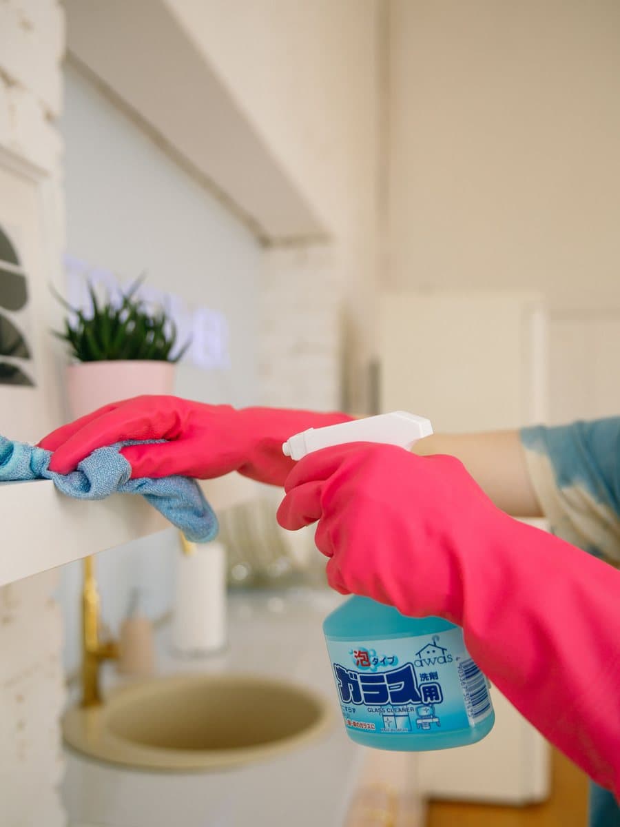 Gloved hands wiping down a bathroom shelf with glass cleaner