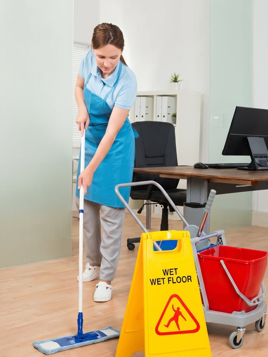 Cleaner mopping an office floor beside a wet floor sign and cart